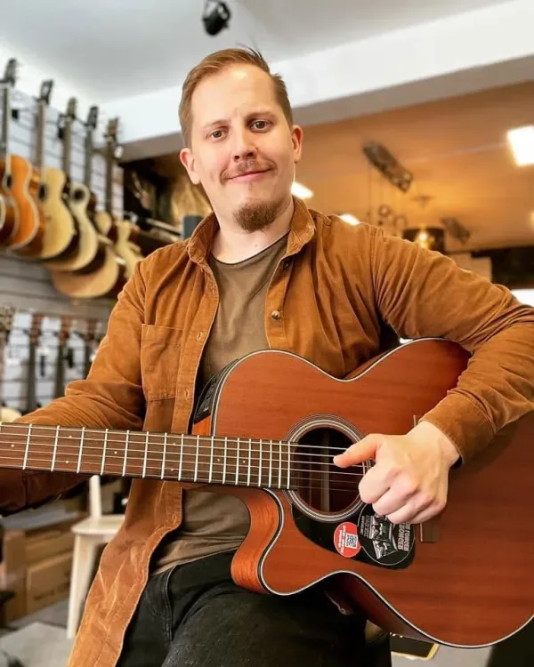 Mann mit Gitarre in Musikladen, lächelt in die Kamera, Gitarren im Hintergrund.